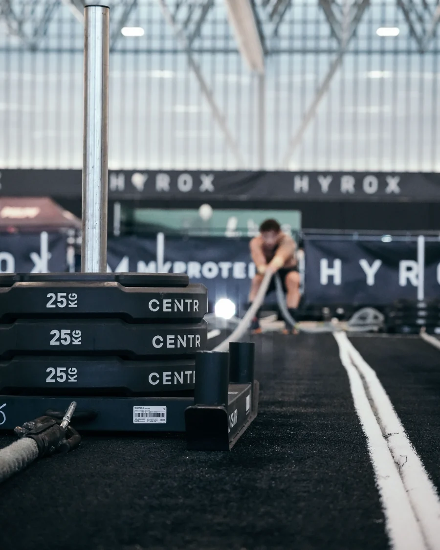 Aire de compétition Hyrox : sled chargé au premier plan, athlète au battle rope